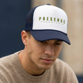 Man wearing a Presence Essentials trucker hat with white foam front panel and navy mesh back in a casual outdoor setting.
